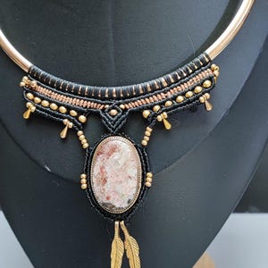 May include: A handcrafted necklace featuring a gold-tone metal collar and a central pendant. The pendant has an oval, speckled stone in a gold setting, with black macrame detailing and gold accents. Two gold leaf charms dangle below.