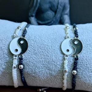May include: Two Yin Yang bracelets. Each bracelet has a white and black Yin Yang symbol. The bracelets are on a grey surface, with a Buddha statue in the background. The bracelets are made of white and black cord.