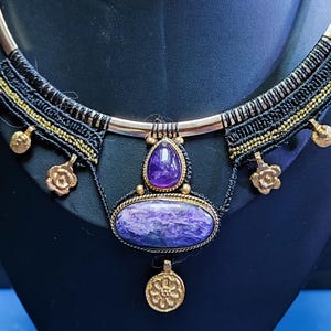 May include: A handcrafted necklace featuring a large, oval purple stone pendant with a smaller, teardrop-shaped stone above it. The necklace has a black and gold woven band with decorative gold accents and a gold-colored metal frame.