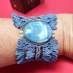Puede incluir: Pulsera de macram&eacute; azul con una piedra ovalada grande de color azul claro engastada en un marco de metal dorado. La pulsera est&aacute; en una mu&ntilde;eca sobre un fondo rojo. Una geoda de cristal est&aacute; en el fondo.