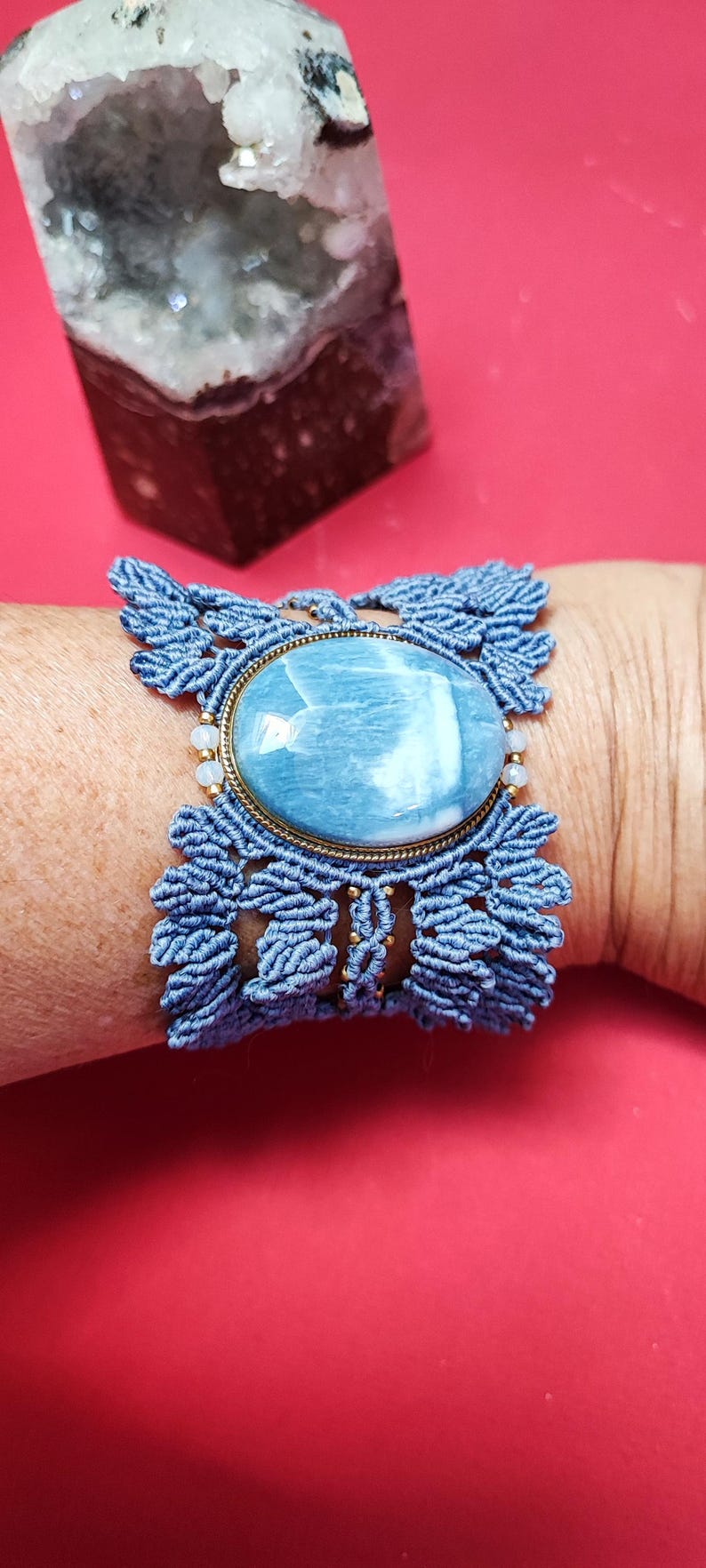 Puede incluir: Pulsera de macram&eacute; azul con una piedra ovalada grande de color azul claro engastada en un marco de metal dorado. La pulsera est&aacute; en una mu&ntilde;eca sobre un fondo rojo. Una geoda de cristal est&aacute; en el fondo.