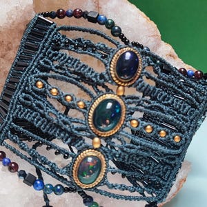 Könnte beinhalten: Ein dunkelblaugrünes Makramee-Armband mit drei ovalen Cabochon-Steinen in goldfarbenen Fassungen. Das geflochtene Kordeldesign ist mit kleinen goldenen Perlen, schwarzen zylindrischen Perlen sowie dunkelblauen, roten und grünen Kugeln verziert.