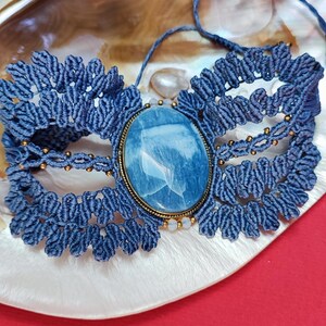Puede incluir: Un collar gargantilla de macram&eacute; azul hecho a mano con una gran piedra preciosa ovalada azul engastada en un marco de metal dorado. El collar se exhibe sobre una concha blanca y dorada sobre un fondo rojo.