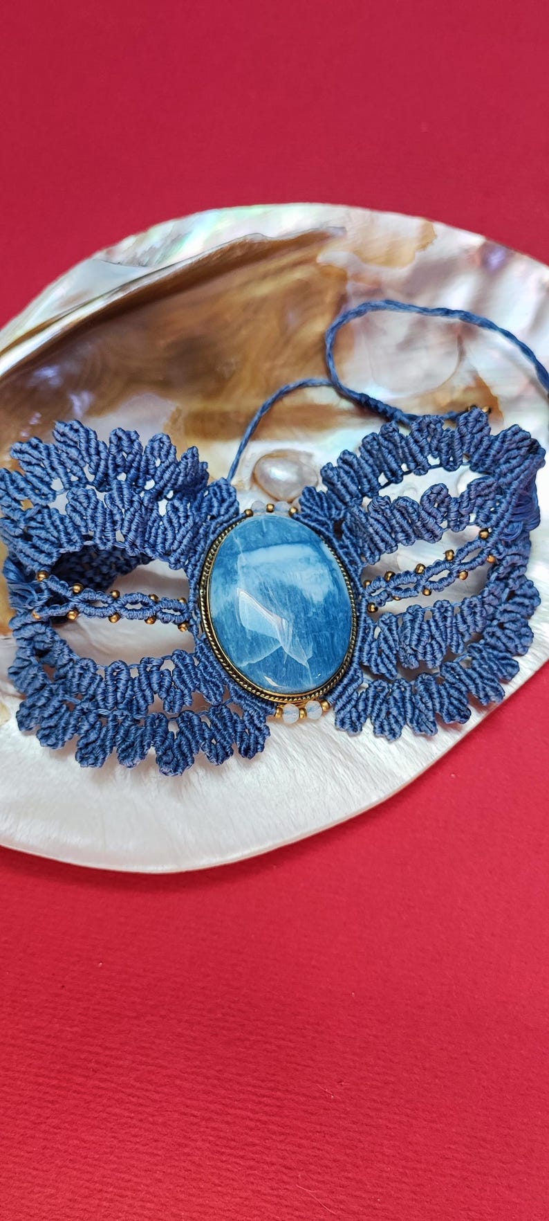 Puede incluir: Un collar gargantilla de macram&eacute; azul hecho a mano con una gran piedra preciosa ovalada azul engastada en un marco de metal dorado. El collar se exhibe sobre una concha blanca y dorada sobre un fondo rojo.