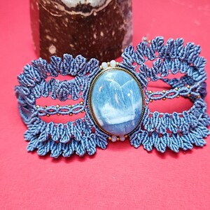 Puede incluir: Una pulsera de macram&eacute; azul con una piedra grande, ovalada, azul claro engastada en un marco color lat&oacute;n. La pulsera est&aacute; sobre una superficie roja, con un cristal en el fondo.
