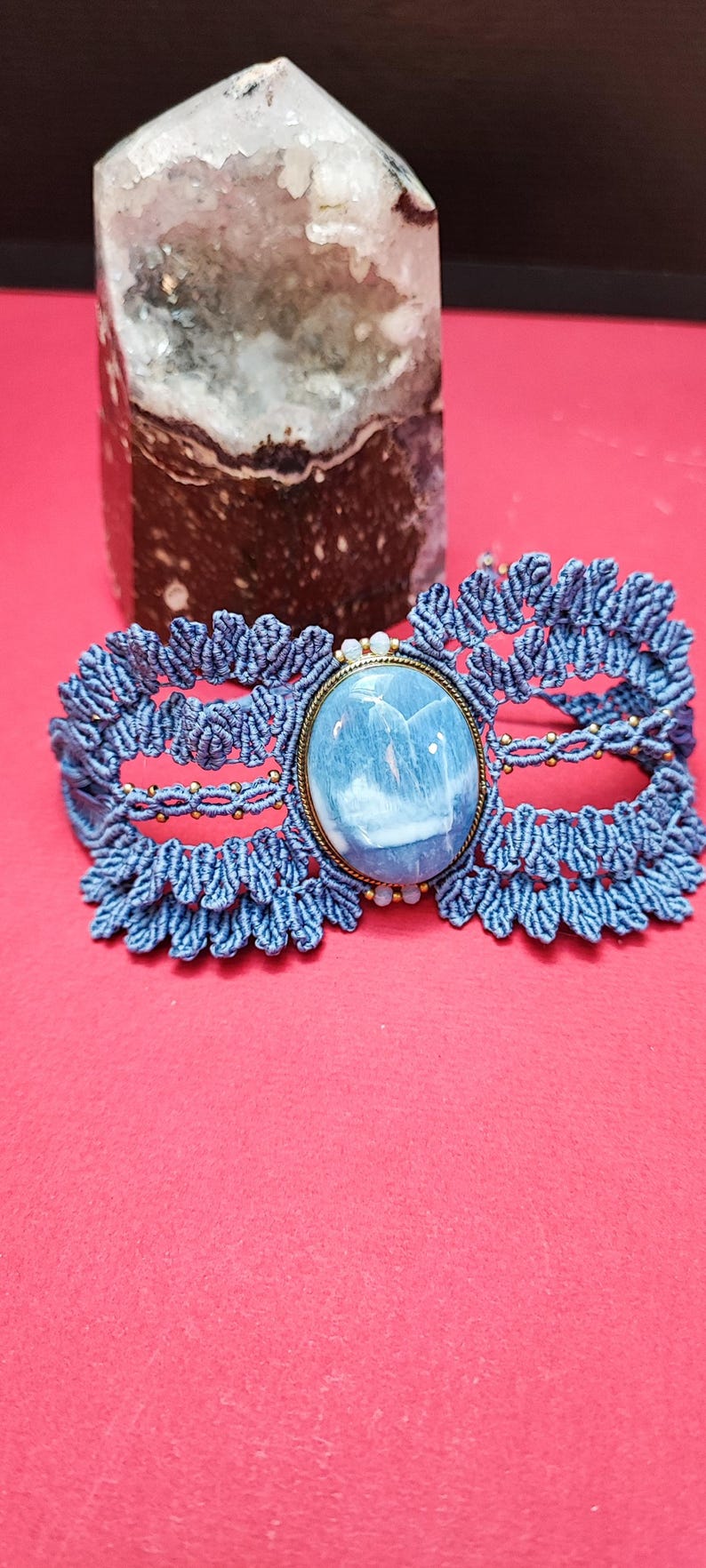 Puede incluir: Una pulsera de macram&eacute; azul con una piedra grande, ovalada, azul claro engastada en un marco color lat&oacute;n. La pulsera est&aacute; sobre una superficie roja, con un cristal en el fondo.