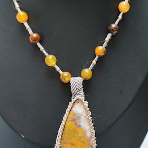 Peut inclure: Un collier avec un pendentif en forme de larme orné d'une pierre d'agate jaune et blanche. Le collier est réalisé avec un cordon gris et de petites perles dorées et ambrées.