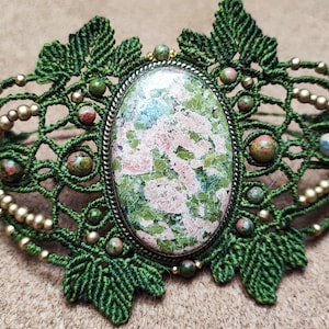 Può includere: Bracciale in macramè fatto a mano con una grande pietra ovale con motivi rosa e verdi, incastonata in una cornice di metallo. Il bracciale è adornato con foglie di macramè verdi, perline dorate e piccoli accenti di pietra.