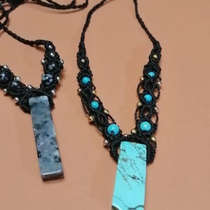 Peut inclure: Deux colliers avec un cordon noir et des pendentifs en pierres précieuses. L'un a un pendentif en turquoise bleu et l'autre un pendentif en pierre grise. Les deux colliers ont de petites perles bleues et dorées.