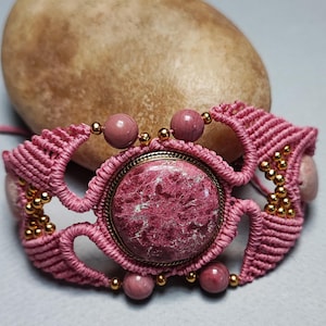 May include: Pink macrame bracelet with a large pink and white stone cabochon in the center. The bracelet is accented with gold beads and smaller pink stones.