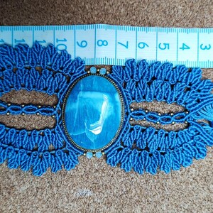 Puede incluir: Un collar gargantilla de macram&eacute; azul hecho a mano con una piedra ovalada azul claro. El collar presenta intrincados nudos y detalles decorativos. Una cinta m&eacute;trica muestra la longitud del collar en cent&iacute;metros.