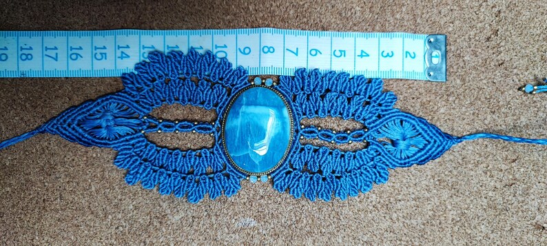 Puede incluir: Un collar gargantilla de macram&eacute; azul hecho a mano con una piedra ovalada azul claro. El collar presenta intrincados nudos y detalles decorativos. Una cinta m&eacute;trica muestra la longitud del collar en cent&iacute;metros.