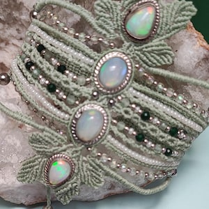 May include: A sage green macrame bracelet featuring three oval-shaped opals set in silver-toned metal. The band is woven with intricate cord patterns, interspersed with rows of small white, clear, and dark green beads, creating a layered texture.