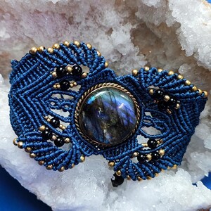 Könnte beinhalten: Ein handgefertigtes Makramee-Armband in Dunkelblau mit einem schillernden Mittelstein. Das Armband ist mit schwarzen und goldenen Perlen verziert, auf einer weißen Kristallgeode und einem blauen Hintergrund.