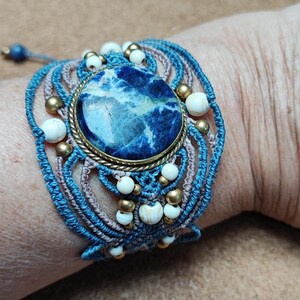 Op de afbeelding: Een macram&eacute; armband met een grote, ovale blauwe steen en witte en gouden accenten. De armband is gemaakt met blauwe en bruine koorden en bevat witte en gouden kralen. De armband zit om een pols.