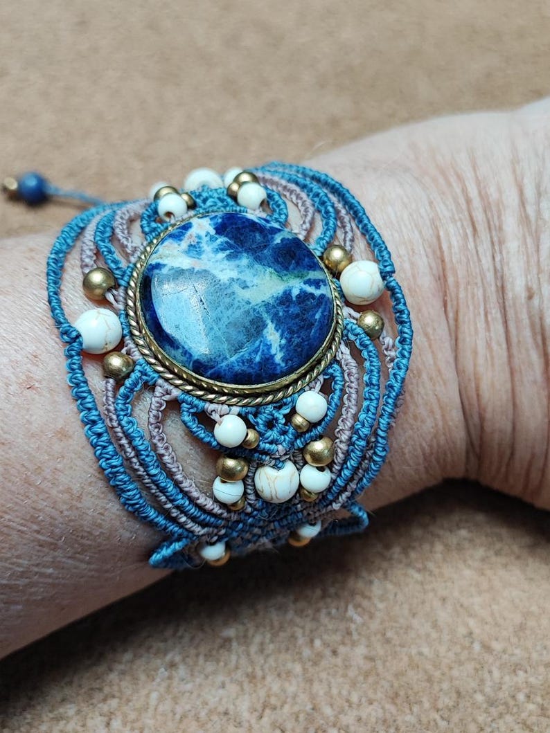 Op de afbeelding: Een macram&eacute; armband met een grote, ovale blauwe steen en witte en gouden accenten. De armband is gemaakt met blauwe en bruine koorden en bevat witte en gouden kralen. De armband zit om een pols.
