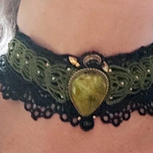 May include: A black macrame choker necklace with a green and gold gemstone pendant. The choker features a green macrame design with small white beads and a black lace trim. The pendant is a teardrop shape.