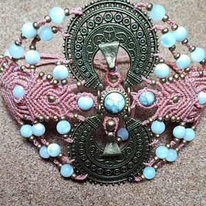 Può includere: Un braccialetto in macramé rosa con perline bianche e un grande ciondolo in metallo dorato decorato.