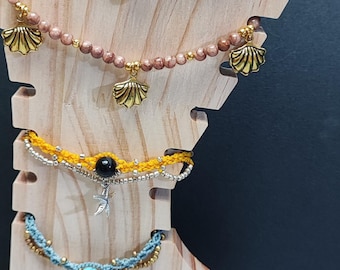 May include: A wooden jewellery display stand with four necklaces and three bracelets. The necklaces feature different coloured beads and charms, including silver teardrop charms, gold shell charms, and a silver starfish charm. The bracelets are made of different coloured cords and beads.