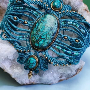 Könnte beinhalten: Türkisfarbenes Makramee-Armband mit einem ovalen türkisfarbenen Mittelstein und kleineren türkisfarbenen Steinen auf jeder Seite. Das Armband besteht aus teal-farbenem Kordel mit goldfarbenen Perlen und Akzenten. Es wird auf einem weißen und grauen Stein präsentiert.