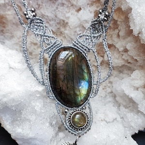 Può includere: Una collana in macramè argentato fatta a mano con una grande pietra di labradorite ovale con lampi di colore iridescente. Una pietra rotonda più piccola e un ciondolo a forma di foglia pendono sotto. La collana è esposta su sfondo bianco.