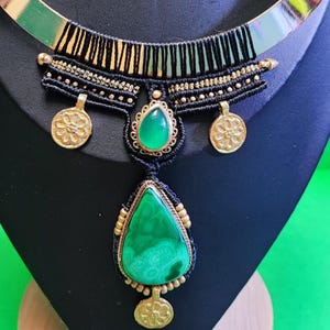 May include: A statement necklace featuring a gold-tone collar and a black macrame design. The necklace has a large, green, teardrop-shaped stone pendant, smaller green stone accents, and gold-tone coin charms. The necklace is displayed on a black bust.
