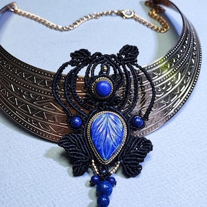 May include: A statement necklace featuring a wide, etched gold-tone collar. The focal point is a black macrame design with a large, carved blue stone pendant and smaller blue stone accents. A delicate gold chain completes the necklace.