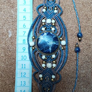 Op de afbeelding: Handgemaakte macram&eacute; bladwijzer met een centrale blauwe steen, witte kralen en messing accenten. De bladwijzer is ongeveer 15 cm lang, met een blauw en bruin geweven ontwerp. Een blauw touwtje met kralen loopt vanaf de bovenkant.