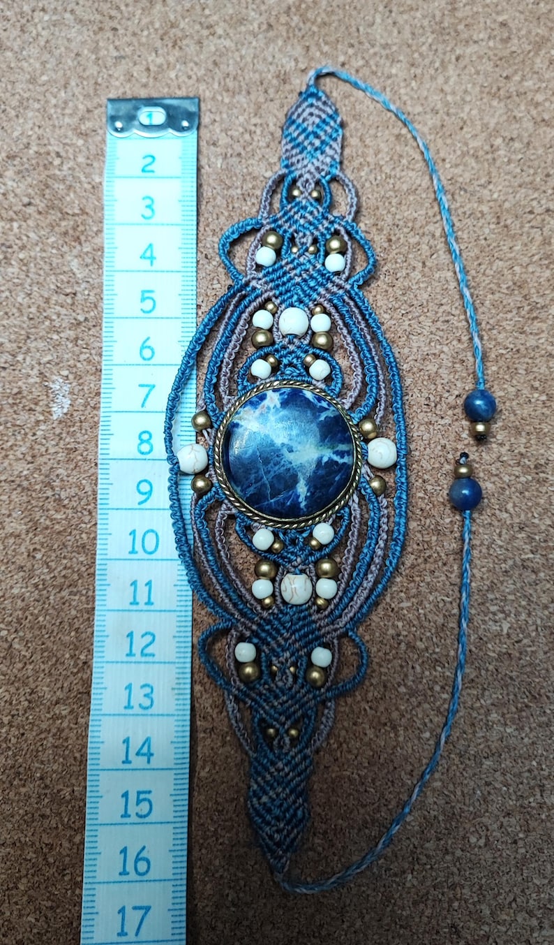 Op de afbeelding: Handgemaakte macram&eacute; bladwijzer met een centrale blauwe steen, witte kralen en messing accenten. De bladwijzer is ongeveer 15 cm lang, met een blauw en bruin geweven ontwerp. Een blauw touwtje met kralen loopt vanaf de bovenkant.