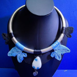 May include: A handcrafted necklace featuring a silver-tone collar with blue and black accents. The necklace has blue leaf-shaped macrame details and a white and blue pendant with a silver leaf charm. Displayed on a black bust against a blue backdrop.