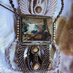 May include: A handcrafted macrame necklace featuring a rectangular pendant with a landscape design. The necklace incorporates beige and brown cords, accented with small, round, beige beads. The pendant is framed in a gold-colored metal.