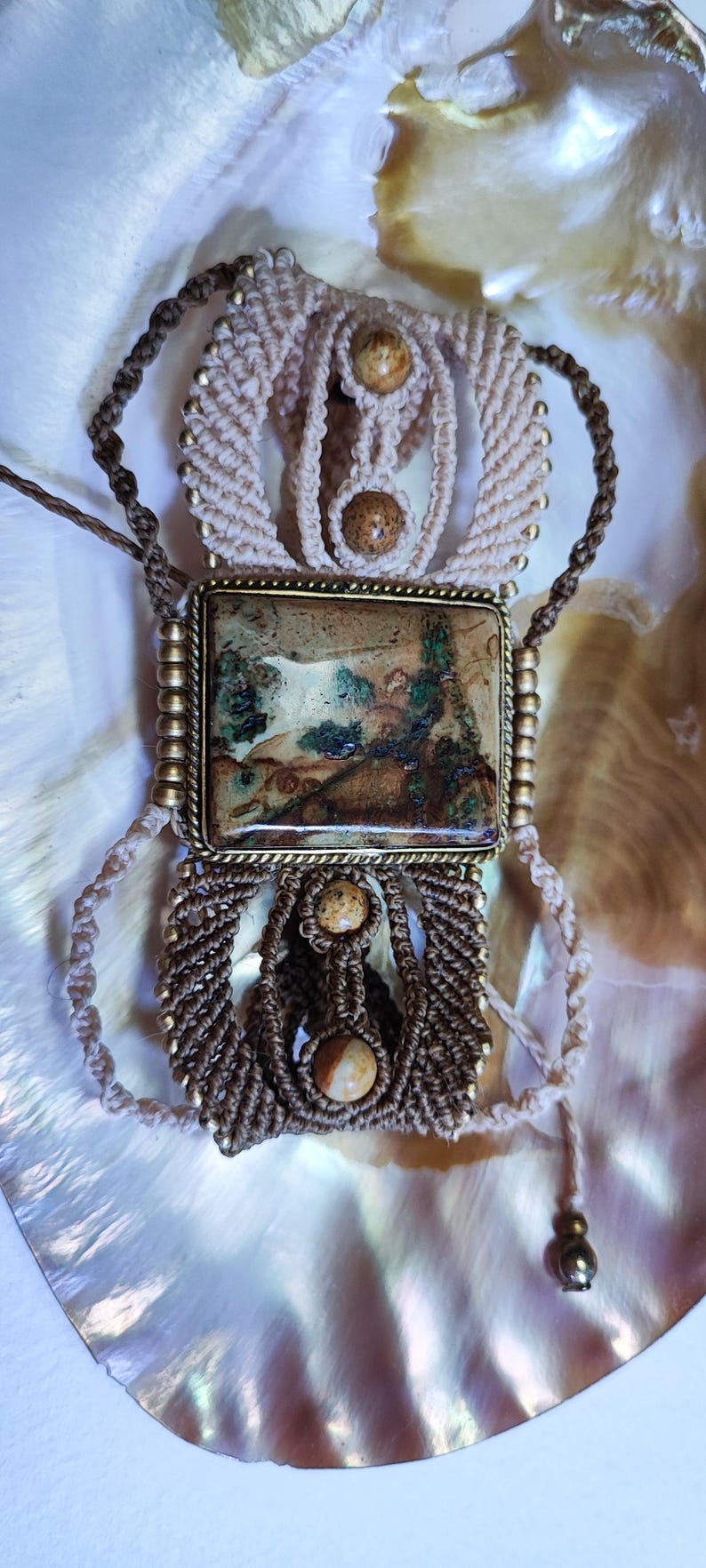 May include: A handcrafted macrame necklace featuring a rectangular pendant with a landscape design. The necklace incorporates beige and brown cords, accented with small, round, beige beads. The pendant is framed in a gold-colored metal.