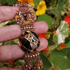 Peut inclure: Un bracelet en macramé marron avec des perles dorées et une grosse perle ronde à motifs noirs et dorés au centre.