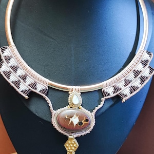 May include: A handcrafted necklace featuring a copper-colored metal collar. The necklace has a central oval stone with a unique pattern, accented by a teardrop-shaped pendant and a decorative gold-colored element. The necklace is displayed on a black bust.