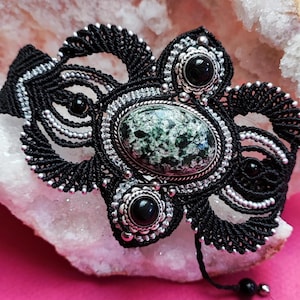 Può includere: Un braccialetto macramè nero fatto a mano con una pietra verde ovale centrale e dettagli argentati. Il braccialetto presenta un'intricata annodatura, piccole perle nere e dettagli in metallo argentato, creando un design unico e artistico.