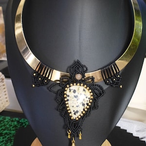 May include: A black and gold choker necklace with a black macrame pendant featuring a large, white and black dalmatian jasper stone.