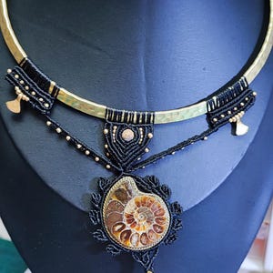 May include: A handcrafted necklace featuring a gold-tone hammered metal collar. The pendant is a fossilized ammonite shell, encased in black macrame with gold accents. The necklace is displayed on a black bust.
