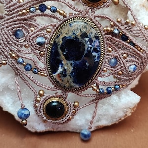 Peut inclure: Un bracelet en macramé avec une grande pierre ovale bleue et crème dans un sertissage en laiton, orné de deux petites pierres noires rondes, de minuscules perles dorées et de perles marbrées bleu et blanc tissées sur un cordon marron clair.
