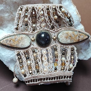 May include: A macrame bracelet crafted from beige cord, featuring a central dark round stone flanked by two elongated, speckled fossilized stone cabochons. The design incorporates intricate woven patterns accented with small brown and cream beads.