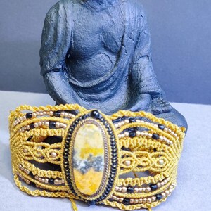 Peut inclure: Bracelet en macramé jaune fait à la main, orné d'une pierre ovale centrale aux tons jaunes et bruns, encadrée de perles noires. Statue de Bouddha en arrière-plan.