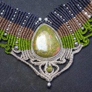 Op de afbeelding: Een groene, bruine en zwarte macramé ketting met een zilveren hanger met een groene en bruine steen.