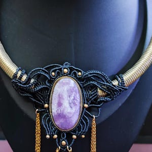 May include: A handcrafted macrame necklace featuring a large, oval, lavender-colored stone pendant. The necklace has a black macrame design with gold accents and a gold chain collar. The pendant is approximately 2 inches long.