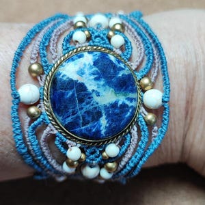 Op de afbeelding: Een handgemaakte macram&eacute; armband met een grote, ronde, blauwe sodaliet steen in het midden. De armband bevat lichtblauwe en mauve macram&eacute; koord, geaccentueerd met witte en goudkleurige kralen. De armband is ongeveer 5 cm breed.