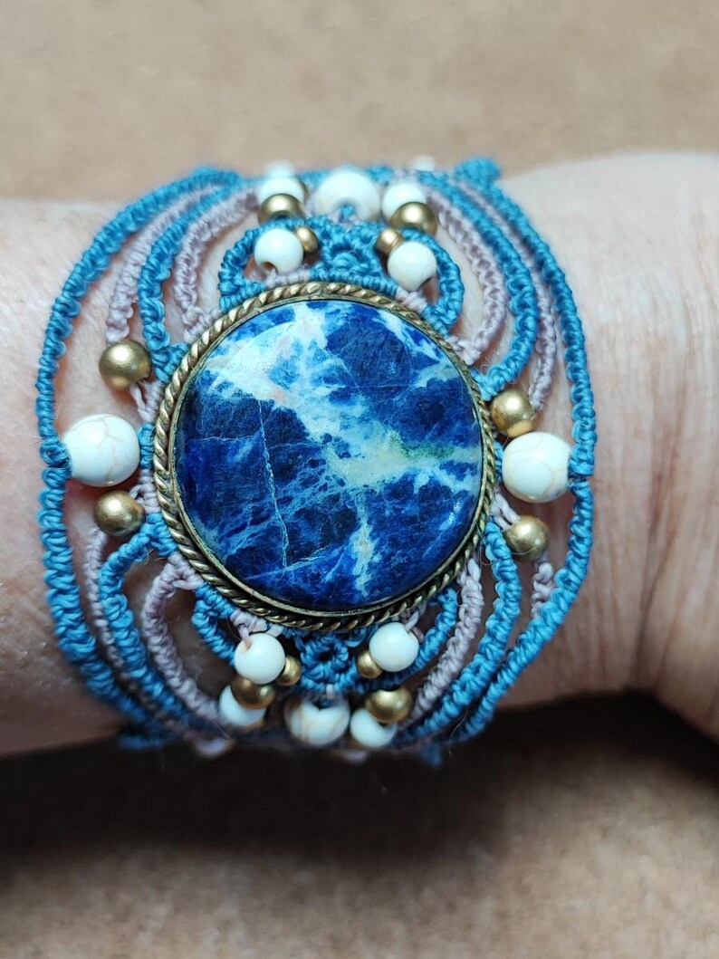Op de afbeelding: Een handgemaakte macram&eacute; armband met een grote, ronde, blauwe sodaliet steen in het midden. De armband bevat lichtblauwe en mauve macram&eacute; koord, geaccentueerd met witte en goudkleurige kralen. De armband is ongeveer 5 cm breed.