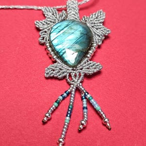 Può includere: Una collana in macramè argentata con un ciondolo in labradorite blu e perline argentate. Il ciondolo è circondato da un design in macramè argentato e ha una catena argentata con una chiusura argentata. La collana ha un fiocco argentato con perline blu e verdi.