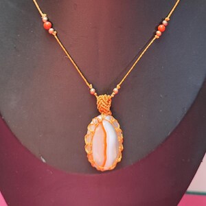 Peut inclure: Collier artisanal avec un pendentif ovale en pierre blanche et orange, serti dans un macramé orange. Le pendentif est suspendu à un cordon orange, orné de petites perles orange et argentées. Fond noir.