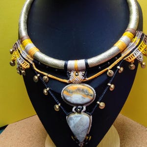 May include: A multi-layered statement necklace featuring a textured gold-tone metal collar, yellow and grey thread wrapping, black cord accents, small gold-tone metal charms, and two large oval and triangular stone pendants with a small elephant charm.