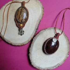 Op de afbeelding: Twee handgemaakte macramé kettingen met bruin koord en ovale stenen hangers. Eén hanger is bruin met witte en gele markeringen, de andere is donkerbruin. Beide hangers hebben kleine metalen accenten.