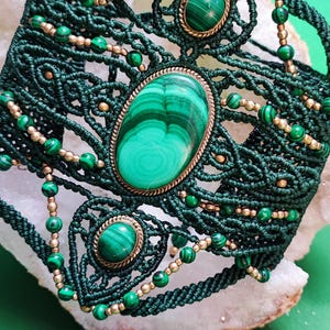 May include: A wide macrame bracelet crafted from dark green cord, featuring three oval malachite cabochons set in brass-toned metal frames. The intricate knotted design is accented with small green malachite beads and metallic gold-toned seed beads.
