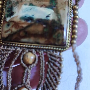 May include: Close-up of a handcrafted macrame bracelet featuring a rectangular stone with brown, green, and white hues set in a gold-colored frame. The bracelet has a beige macrame design with two beige beads.
